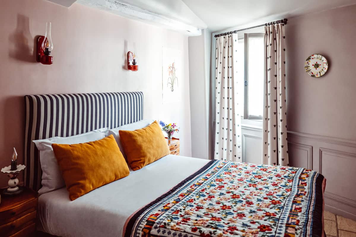 Un lit bien fait, avec une tête de lit à rayures, deux coussins de couleur moutarde et une couvre-lit à motifs floraux. On y trouve une table de chevet avec une lampe et un téléphone. La pièce est dotée d’une fenêtre avec des rideaux à motifs floraux, d’une horloge murale et d’un luminaire fixé au mur.