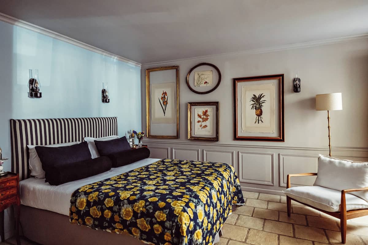 Une chambre bien rangée, avec un grand lit, une chaise et des reproductions encadrées de motifs botaniques accrochées au mur.