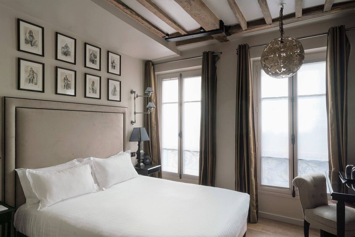 A hotel room with a large bed, multiple framed pictures on the wall, a desk with a lamp, and a view through the window.