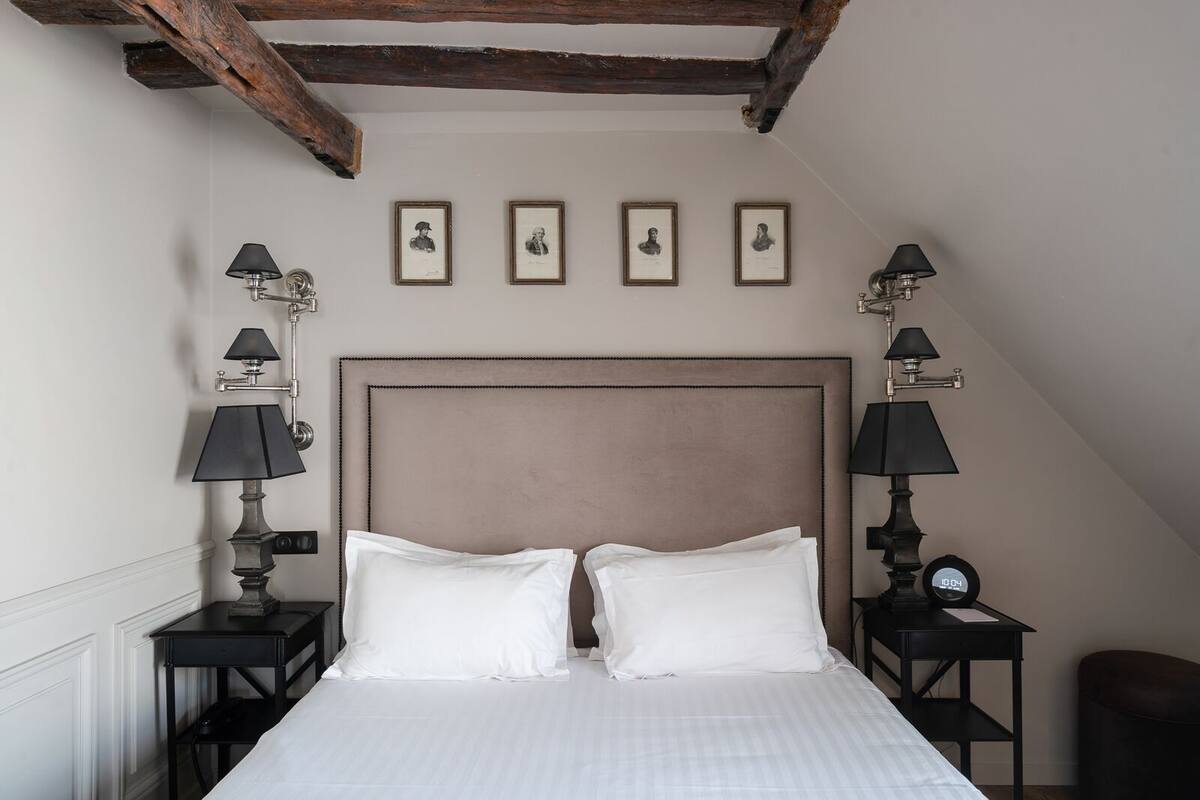A bedroom with a large bed, two bedside tables with lamps, a wooden ceiling beam, and four framed pictures on the wall.