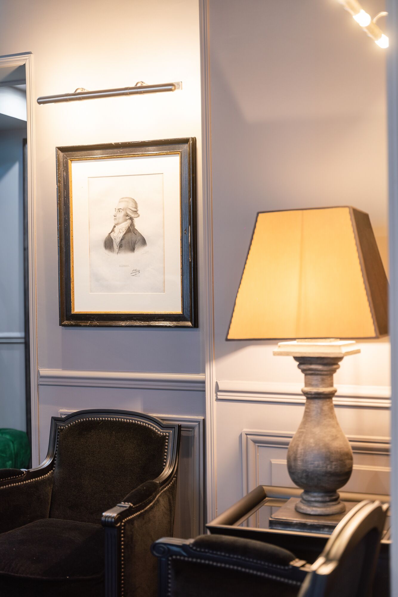 Una habitación con un retrato enmarcado en la pared, una lámpara sobre una mesa y una silla.