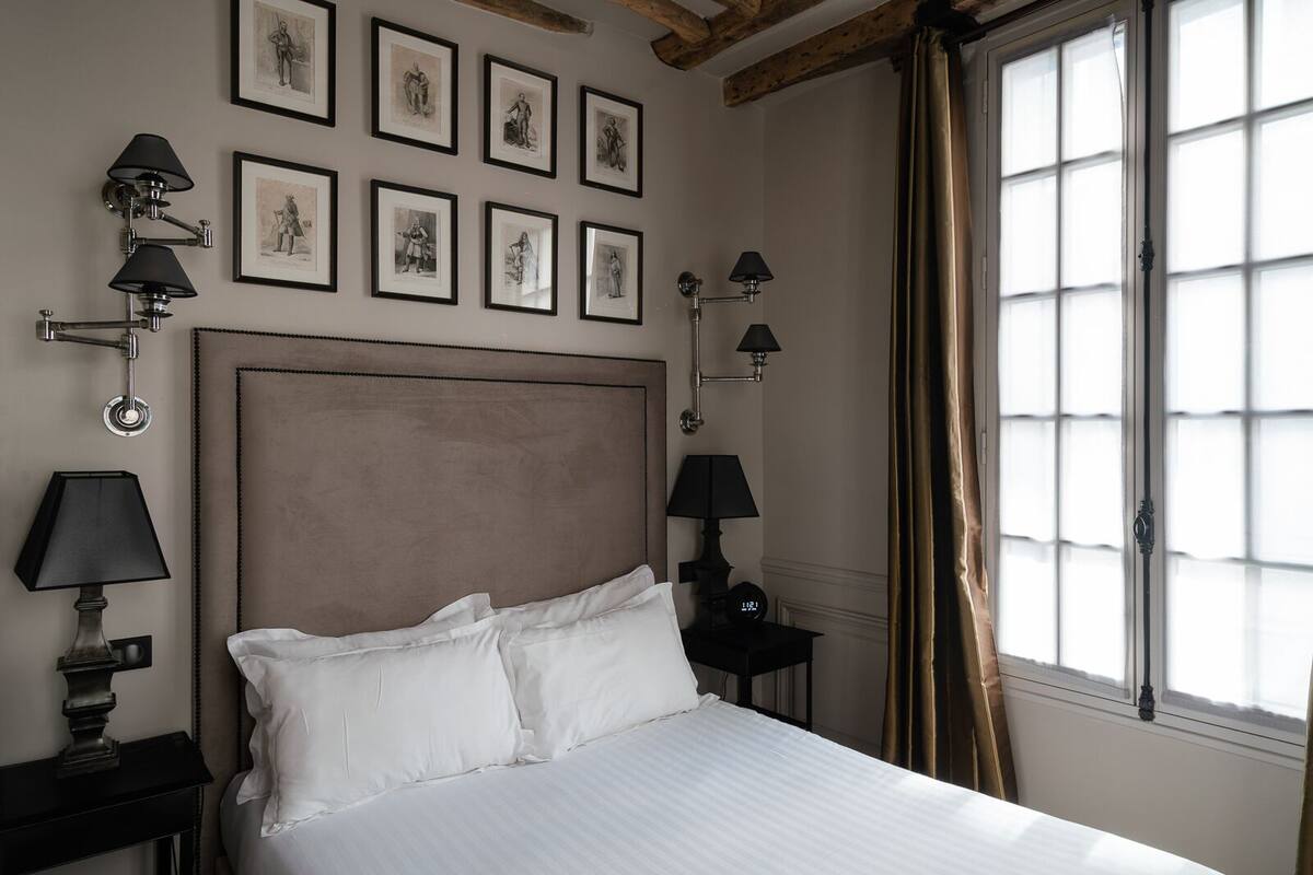 A neatly made bed in a room with a large headboard, multiple framed pictures on the wall, and a window with curtains.