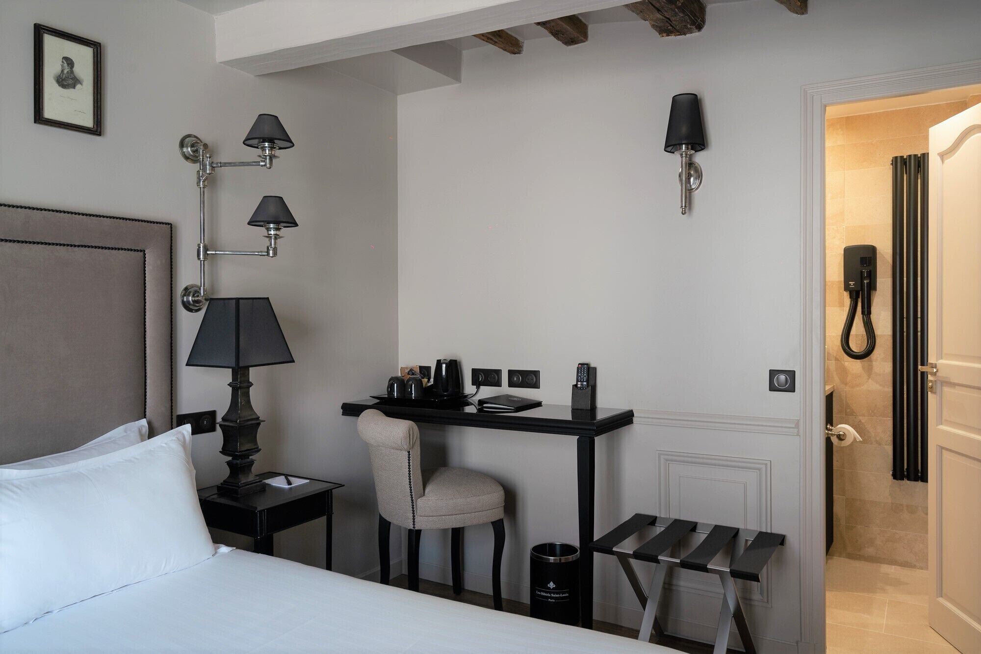 A hotel room with a bed, a desk with a chair, a small table, a trash bin, and a bathroom with a shower.