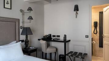 A hotel room with a bed, a desk with a chair, a small table, a trash bin, and a bathroom with a shower.
