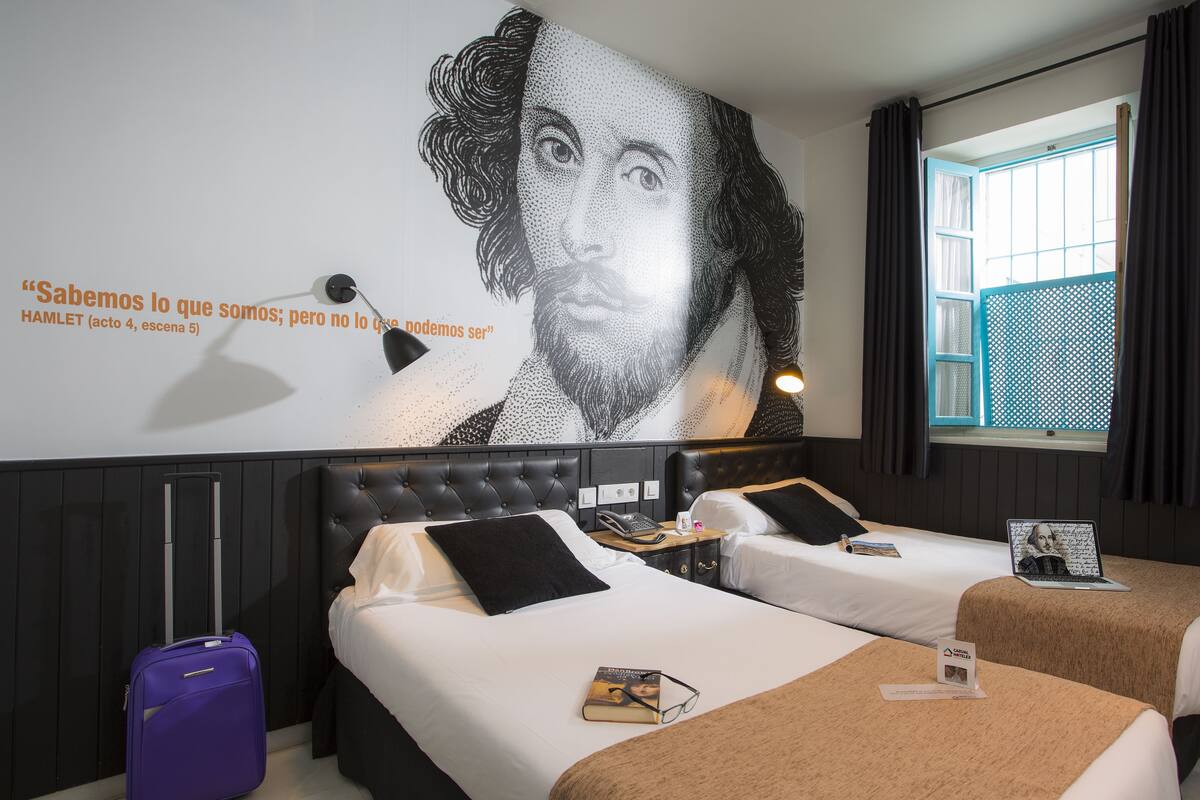Una habitación de hotel con dos camas, un mural grande en la pared, una ventana con cortinas y una maleta morada.