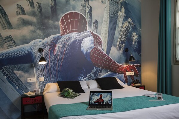 A hotel room with a large Spider-Man mural on the wall, a bed with a laptop, and a nightstand with a lamp and a book.