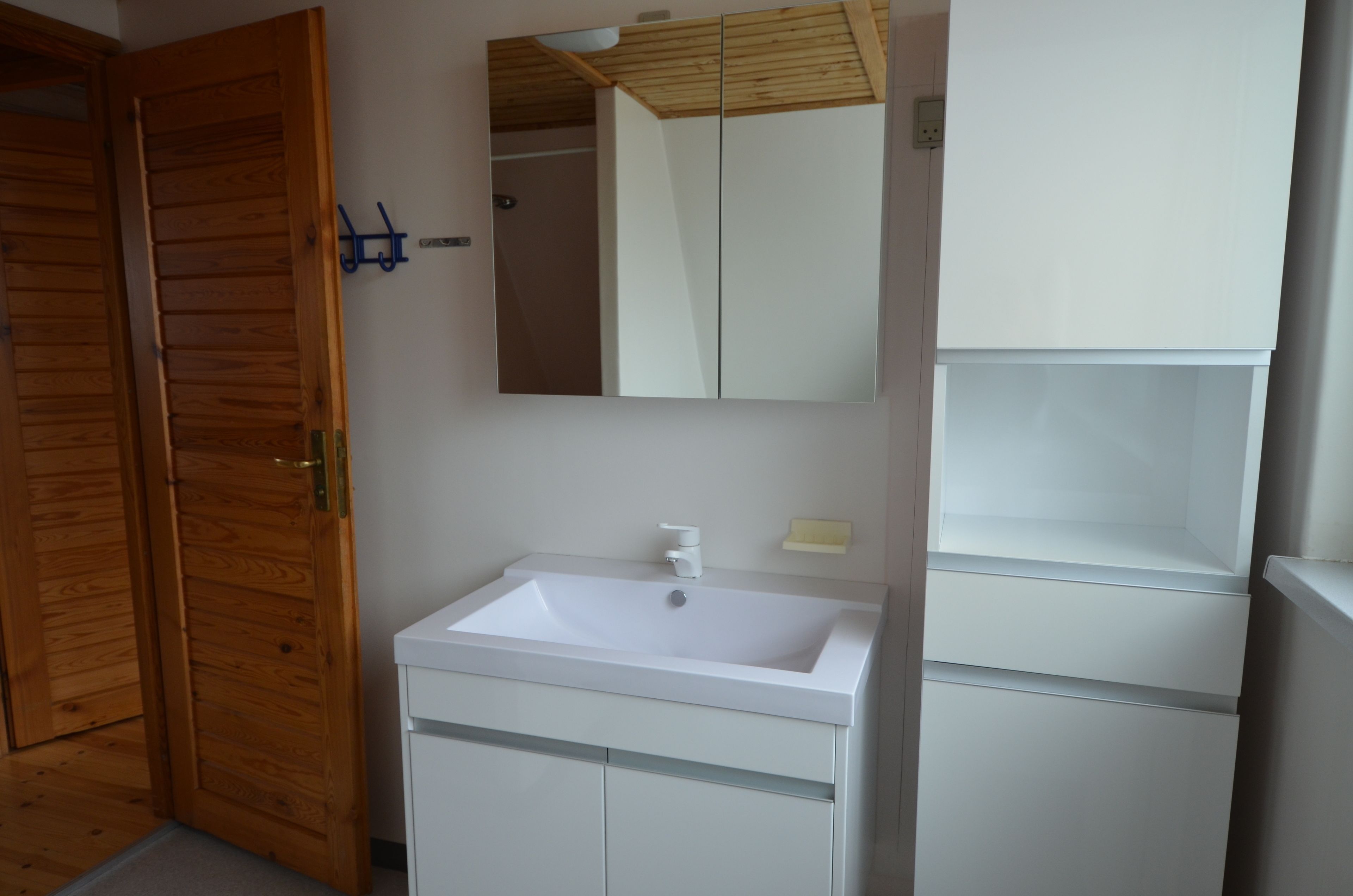 Une salle de bain avec une porte en bois, un lavabo blanc, un miroir et un meuble blanc.