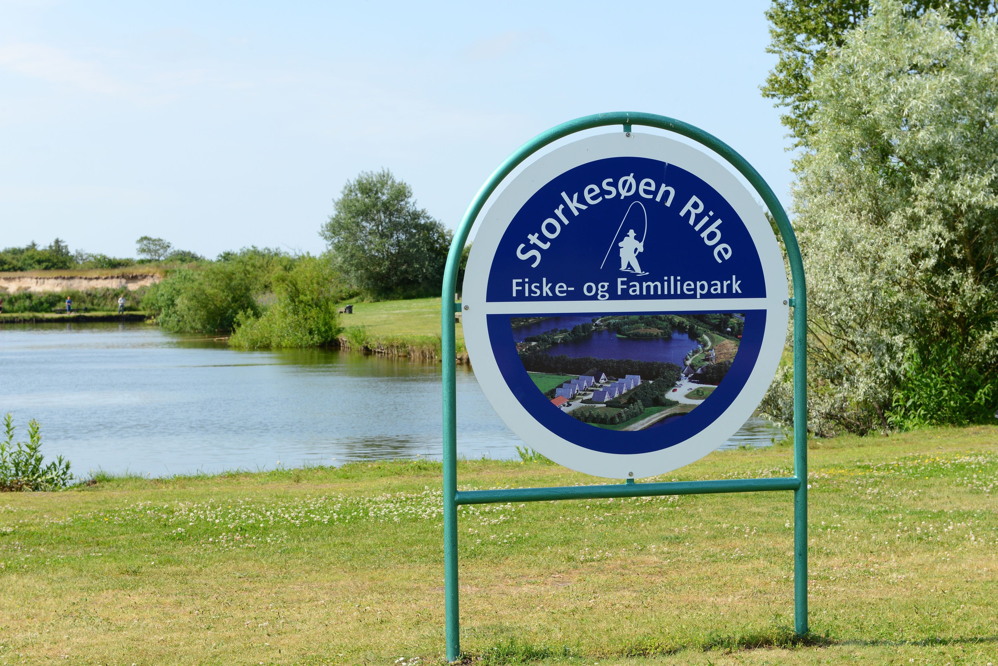 A sign for Storkesøen Ribe, indicating it is a fishing and family park with a view of a lake and greenery.