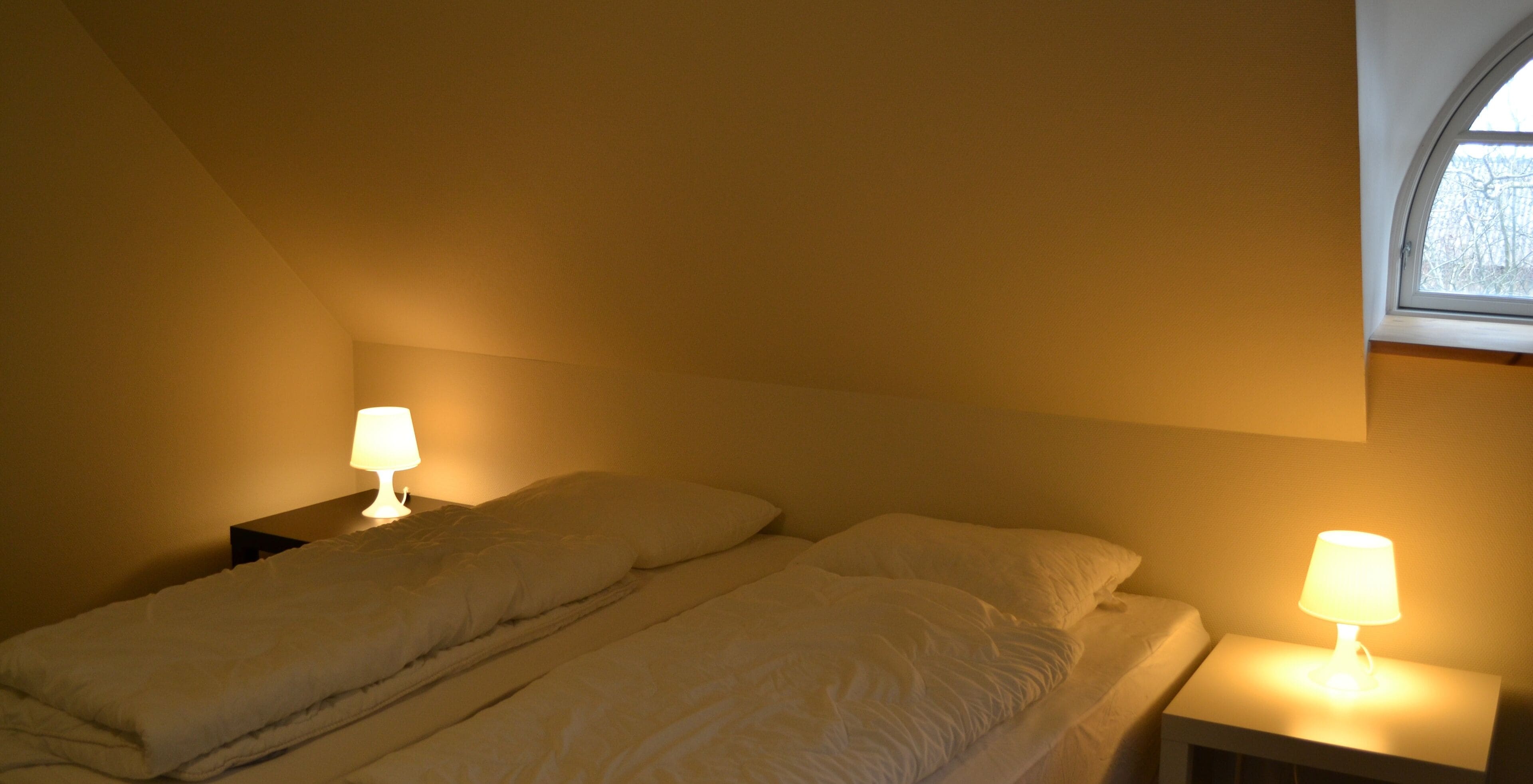 A single bed in a room with a sloped ceiling and a window. There are two bedside tables with lamps.