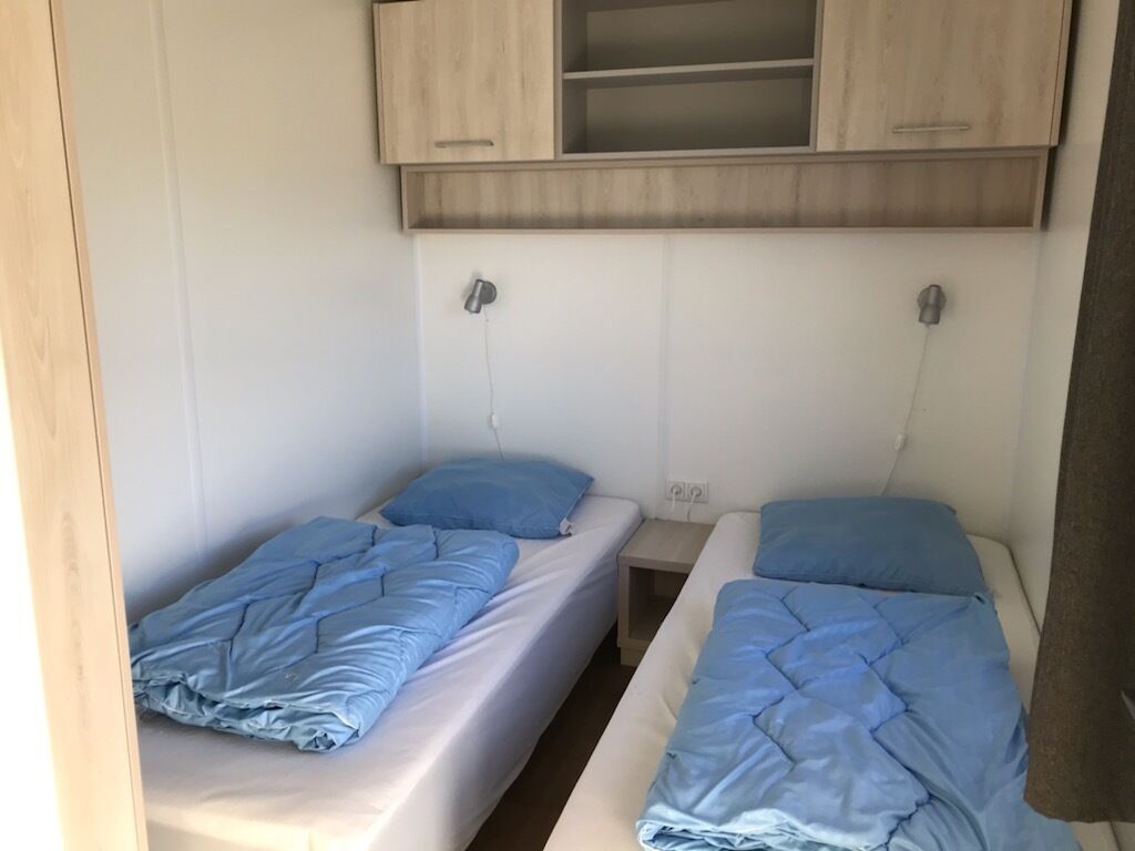 A compact room with two single beds, each with a blue blanket and pillow. There is a wooden cabinet above the beds, and a power outlet is visible on the wall.