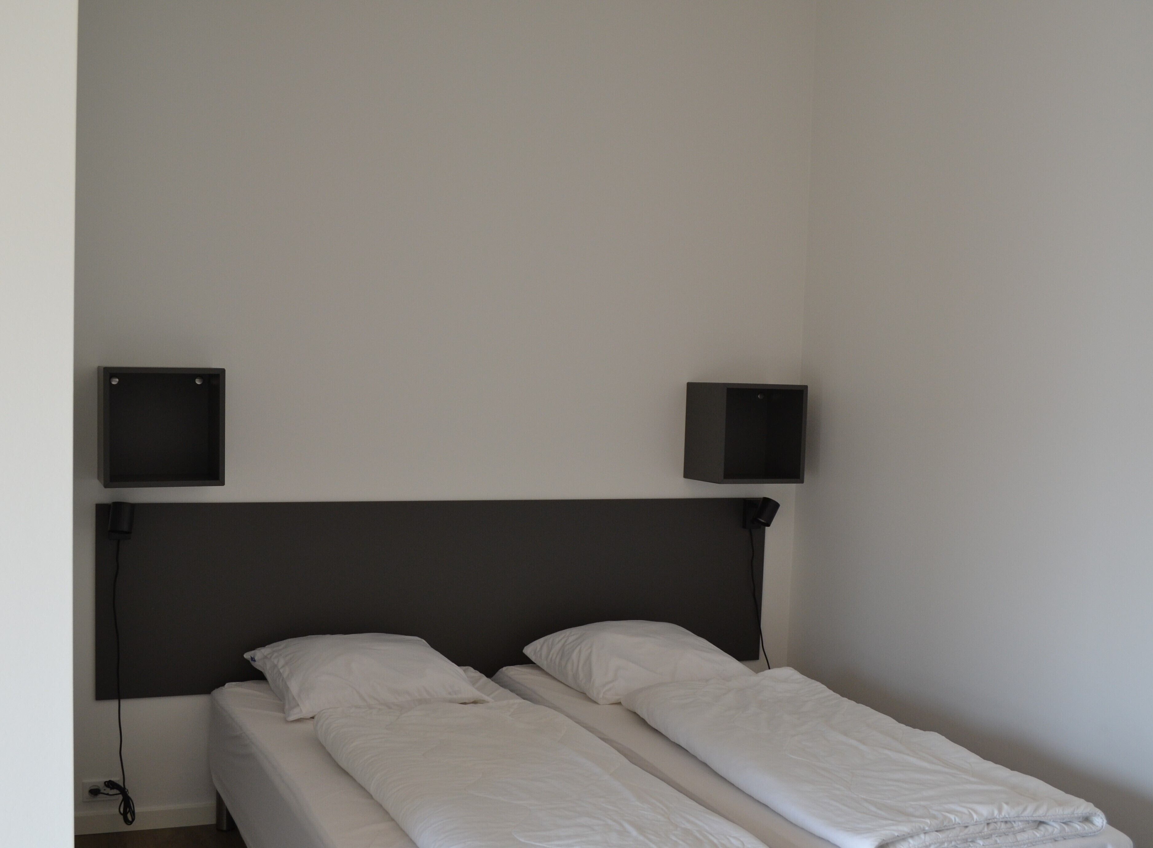 A single bed with a dark headboard, two bedside tables, and a wall-mounted speaker on either side.
