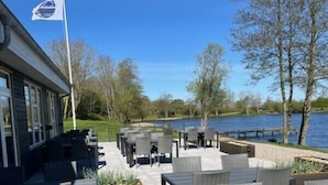An outdoor patio with tables and chairs, overlooking a body of water, with a flag on a flagpole to the left. - Storkesøen Ribe (Ribe)