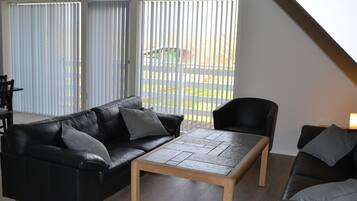 Een moderne woonkamer met een zwarte leren bank, een houten salontafel en een zwarte fauteuil. Grote ramen met zonwering laten natuurlijk licht binnen en bieden uitzicht op de omgeving.
