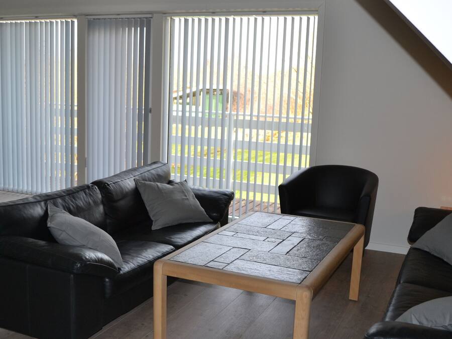 Una sala de estar moderna con un sofá de cuero negro, una mesa de centro de madera y un sillón negro. Amplias ventanas con persianas dejan entrar luz natural y ofrecen vistas al exterior.