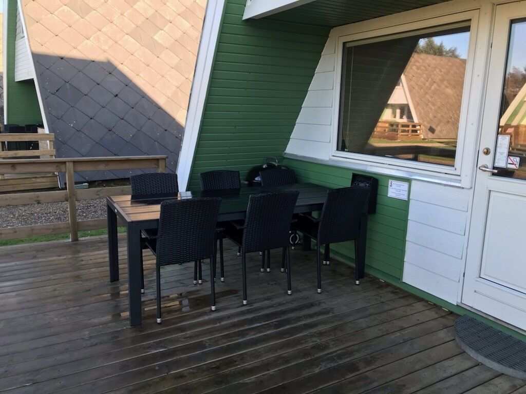 A covered outdoor seating area with a table and chairs, a wooden deck, and a green and white building in the background.