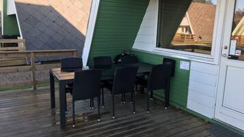 A covered outdoor seating area with a table and chairs, a wooden deck, and a green and white building in the background.