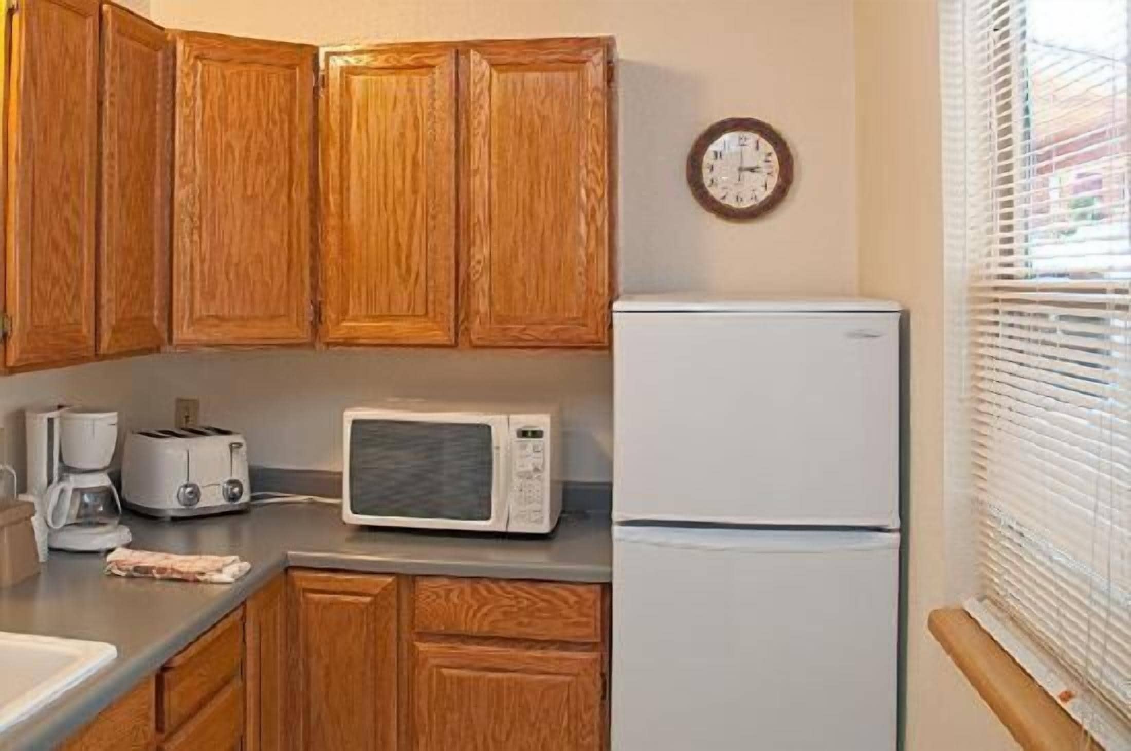 Una cocina con armarios de madera, microondas, tostadora, refrigerador y un reloj en la pared.