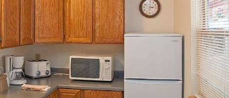 A kitchen with wooden cabinets, a microwave, a toaster, a refrigerator, and a clock on the wall.