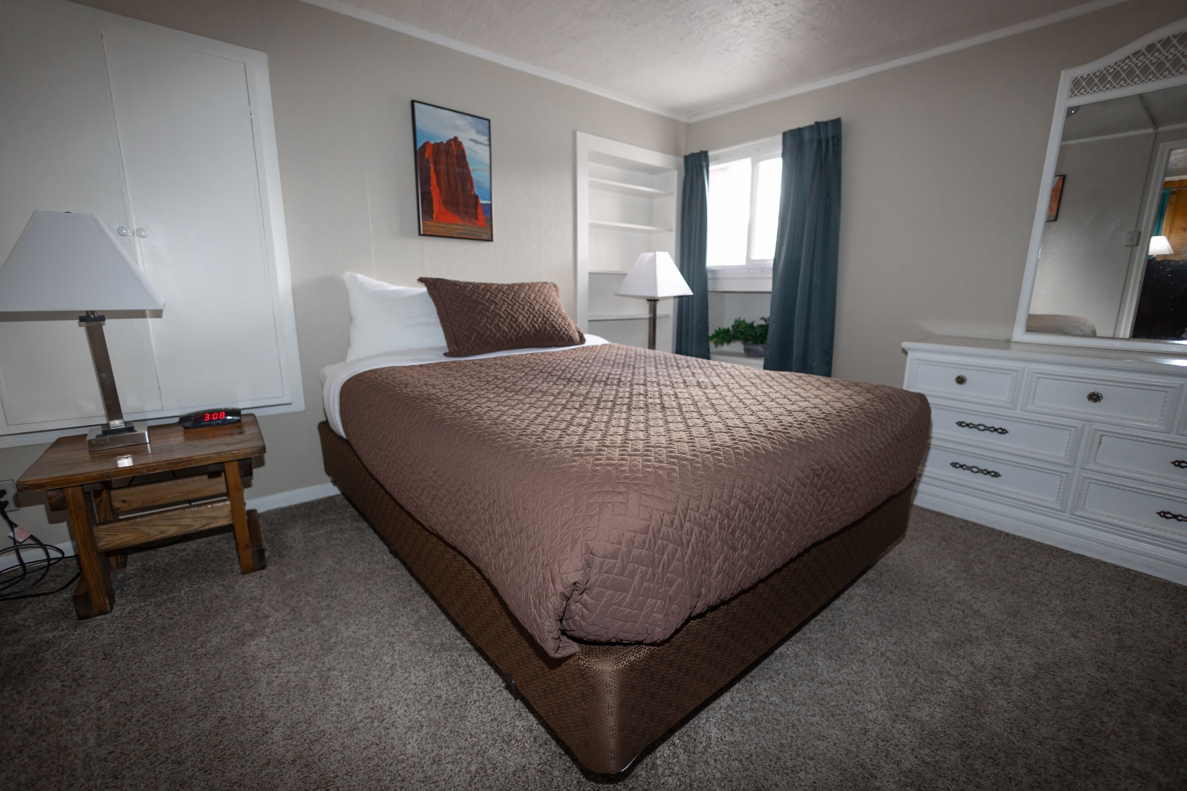 A bedroom with a large bed, a bedside table with a lamp, a dresser, a window with curtains, and a painting on the wall.