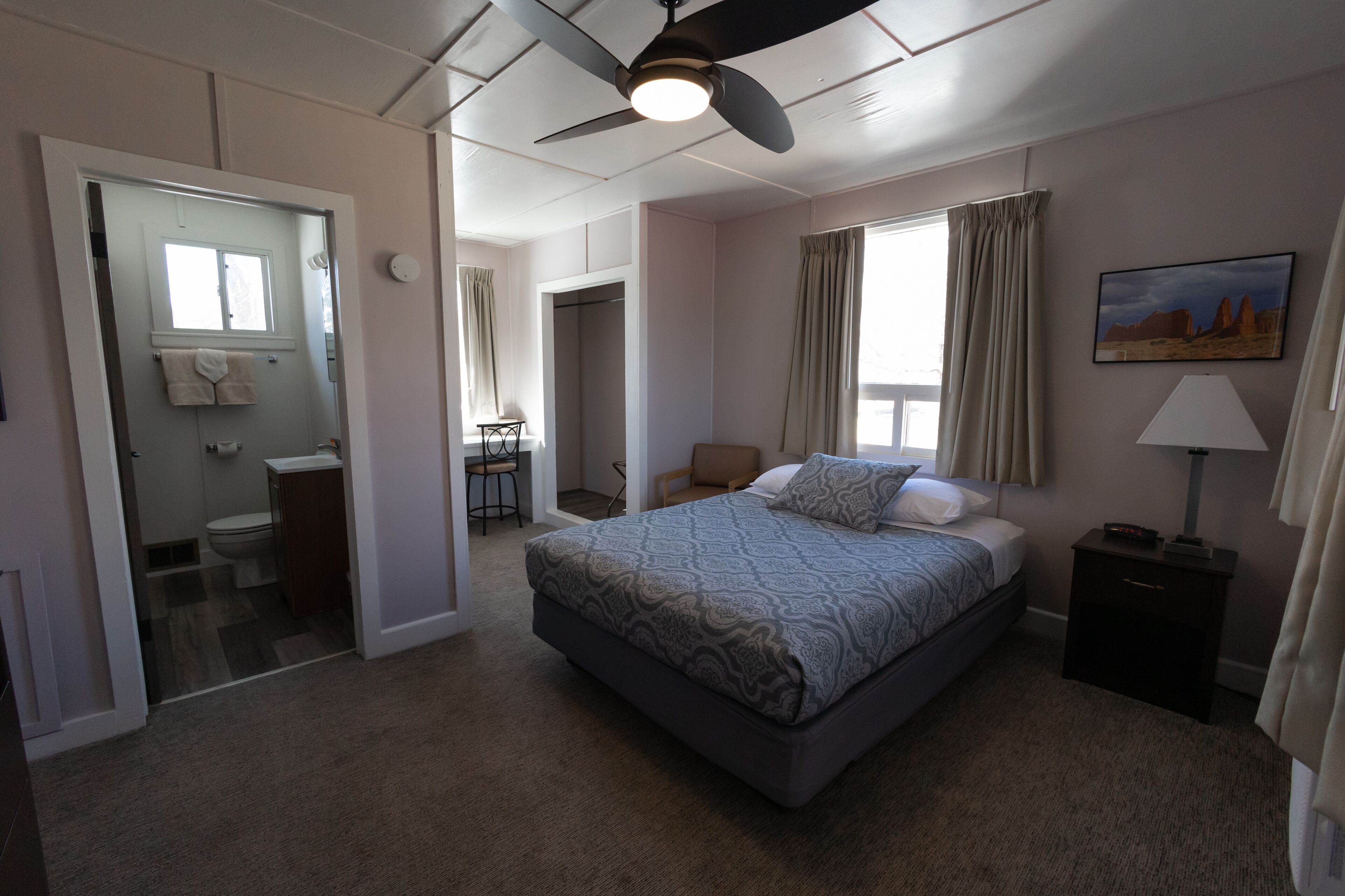 A bedroom with a bed, a ceiling fan, a nightstand, a window with curtains, and a painting on the wall.