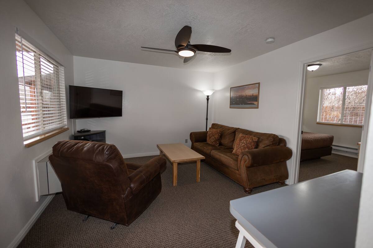 A living room with a brown leather chair, a brown sofa, a wooden coffee table, a ceiling fan, a flat-screen TV, and a painting on the wall.