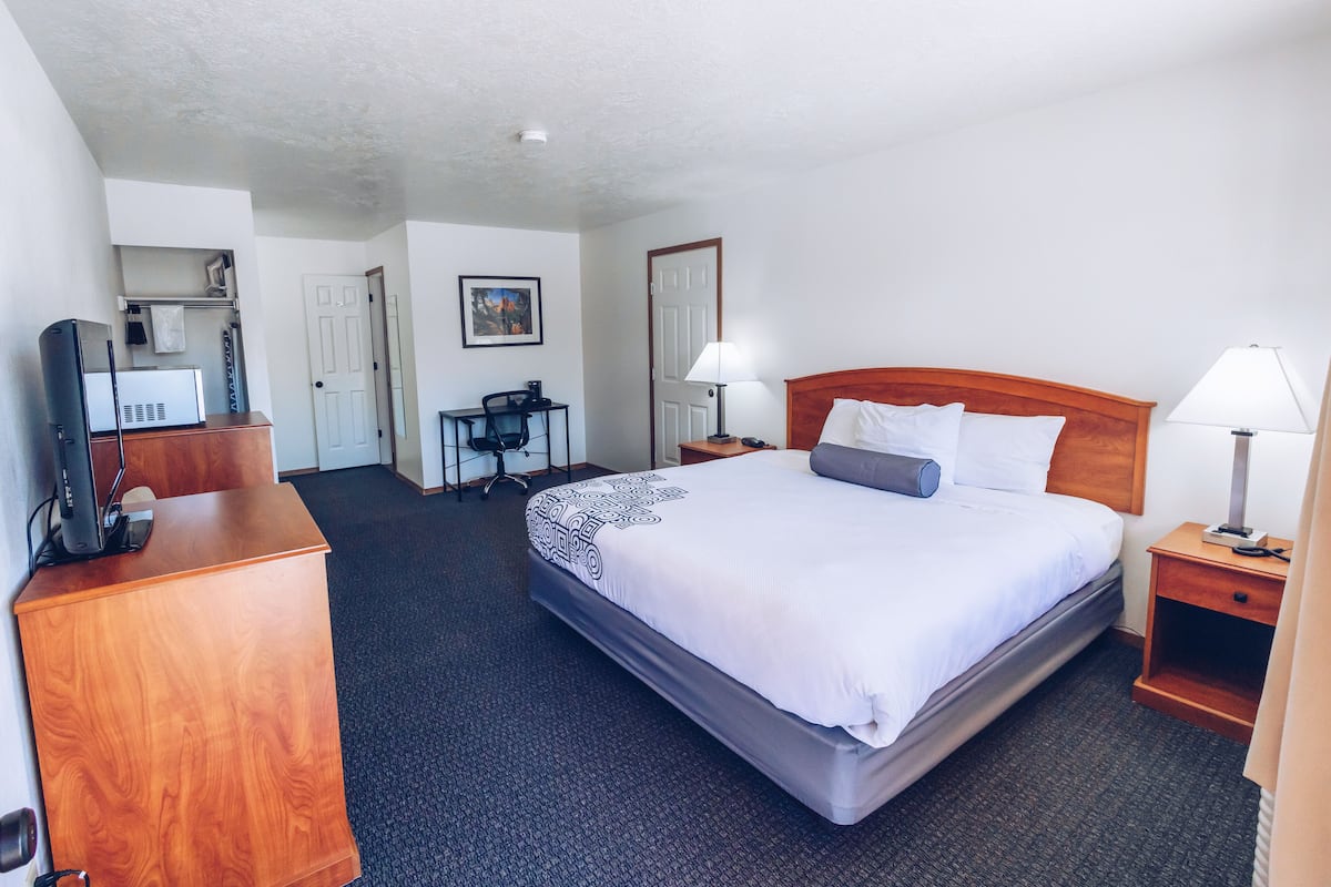 A hotel room with a bed, two bedside tables with lamps, a desk with a chair, a television, and a painting on the wall.