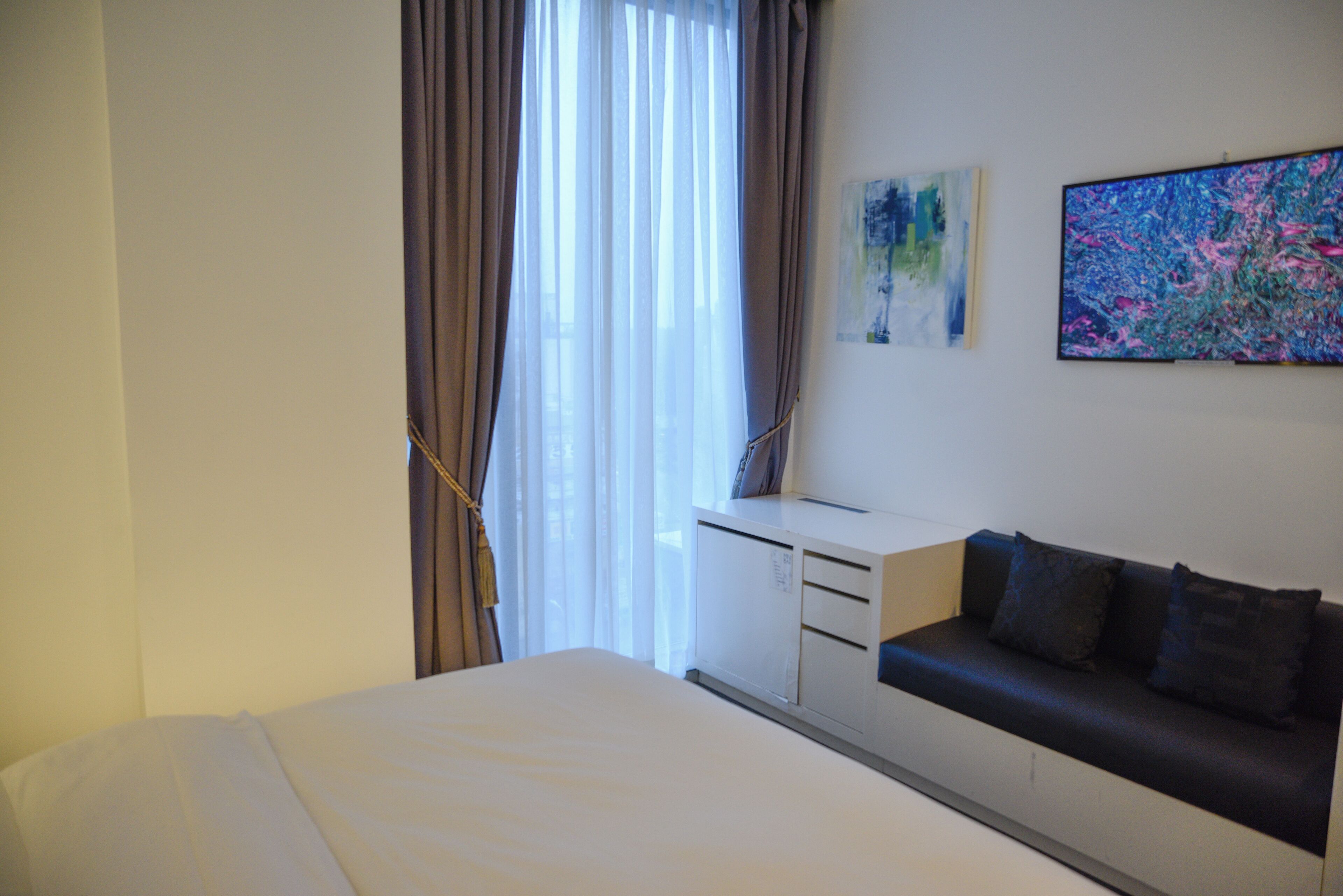 A hotel room with a bed, a desk, a sofa, a window with curtains, and two pieces of wall art.