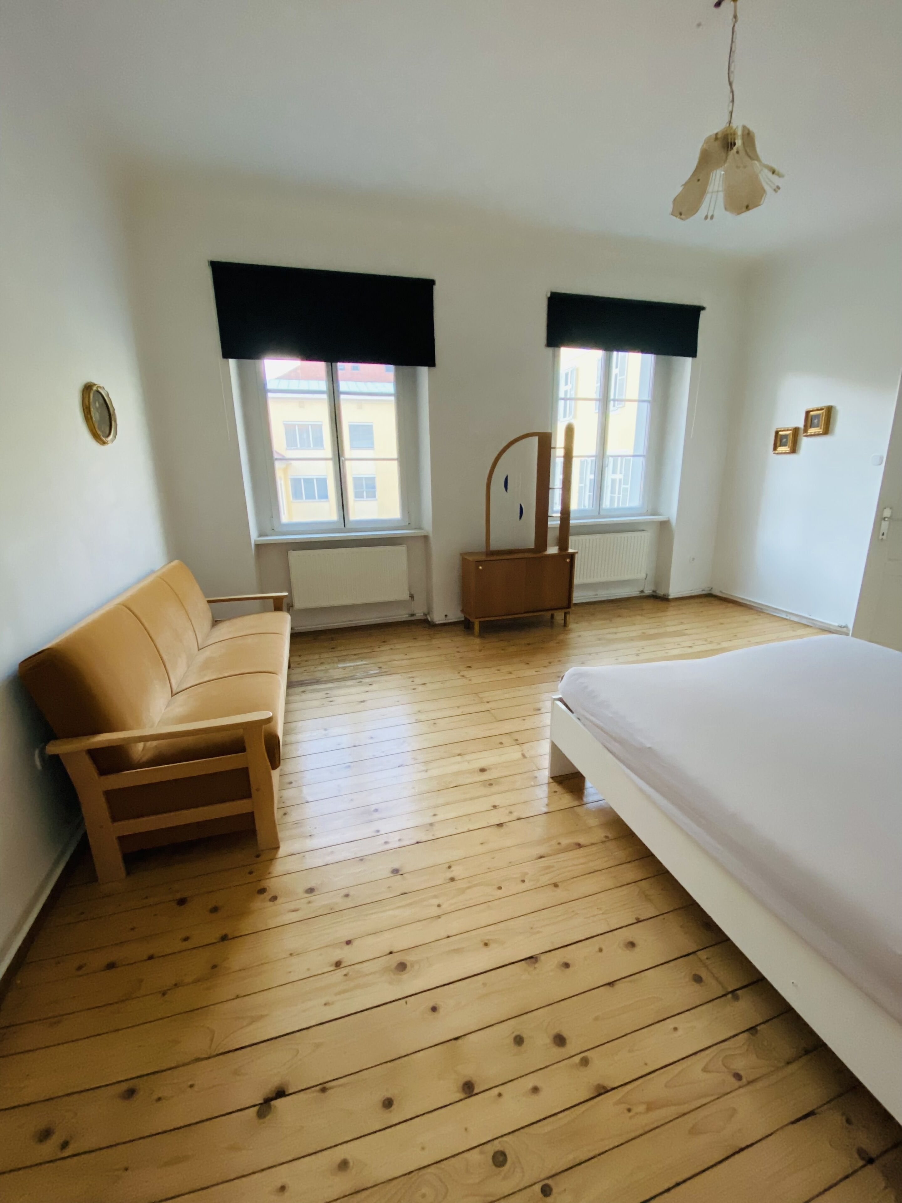 Una habitación con piso de madera, una cama, un sofá, dos ventanas con cortinas negras y una lámpara colgante.