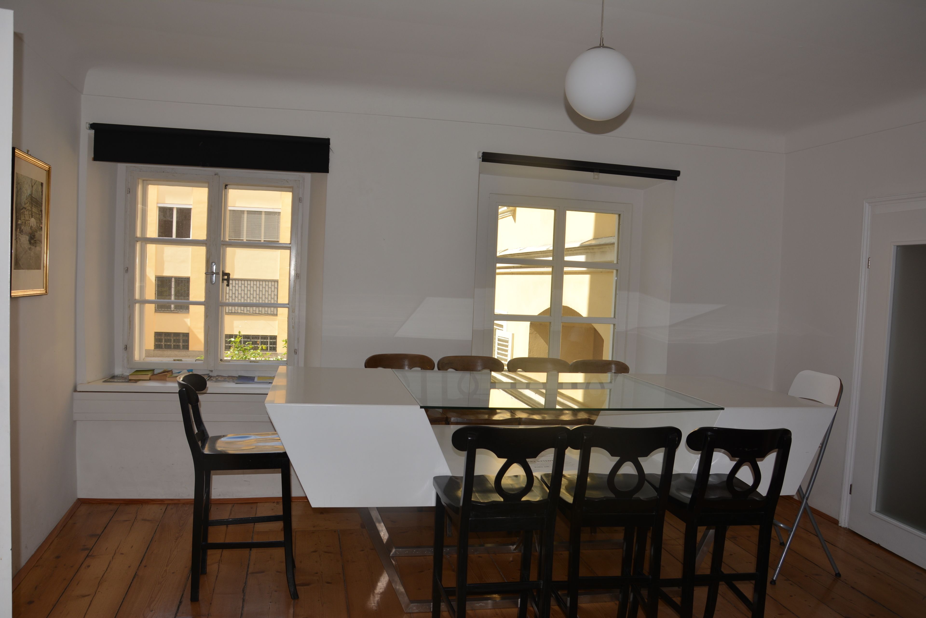 Un comedor con mesa de vidrio y sillas negras, una ventana con persianas y una lámpara colgante.
