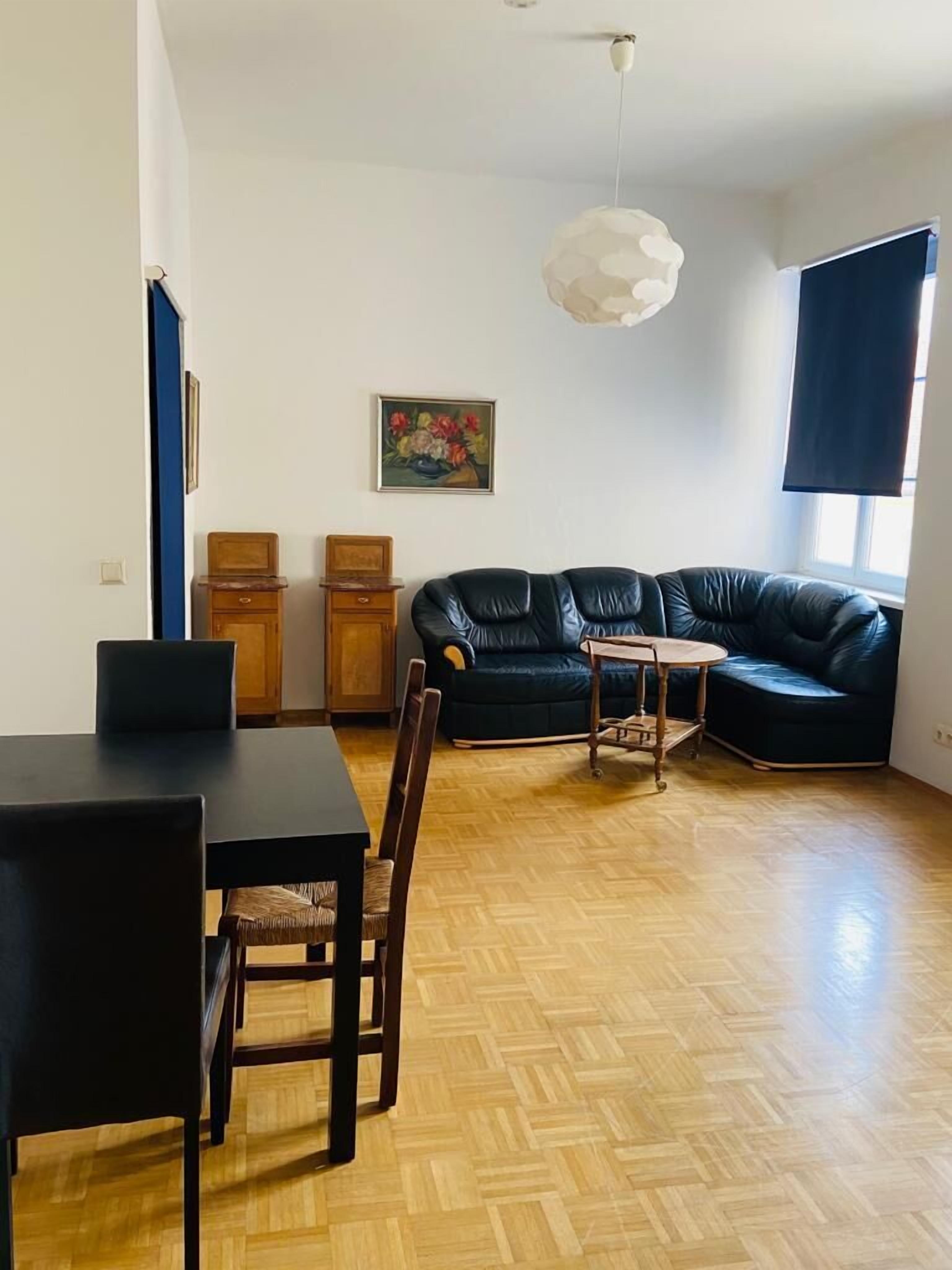 Una sala moderna con un sofá de cuero negro, una mesa de comedor con sillas, una pequeña mesita auxiliar de madera y un colgador de luz.