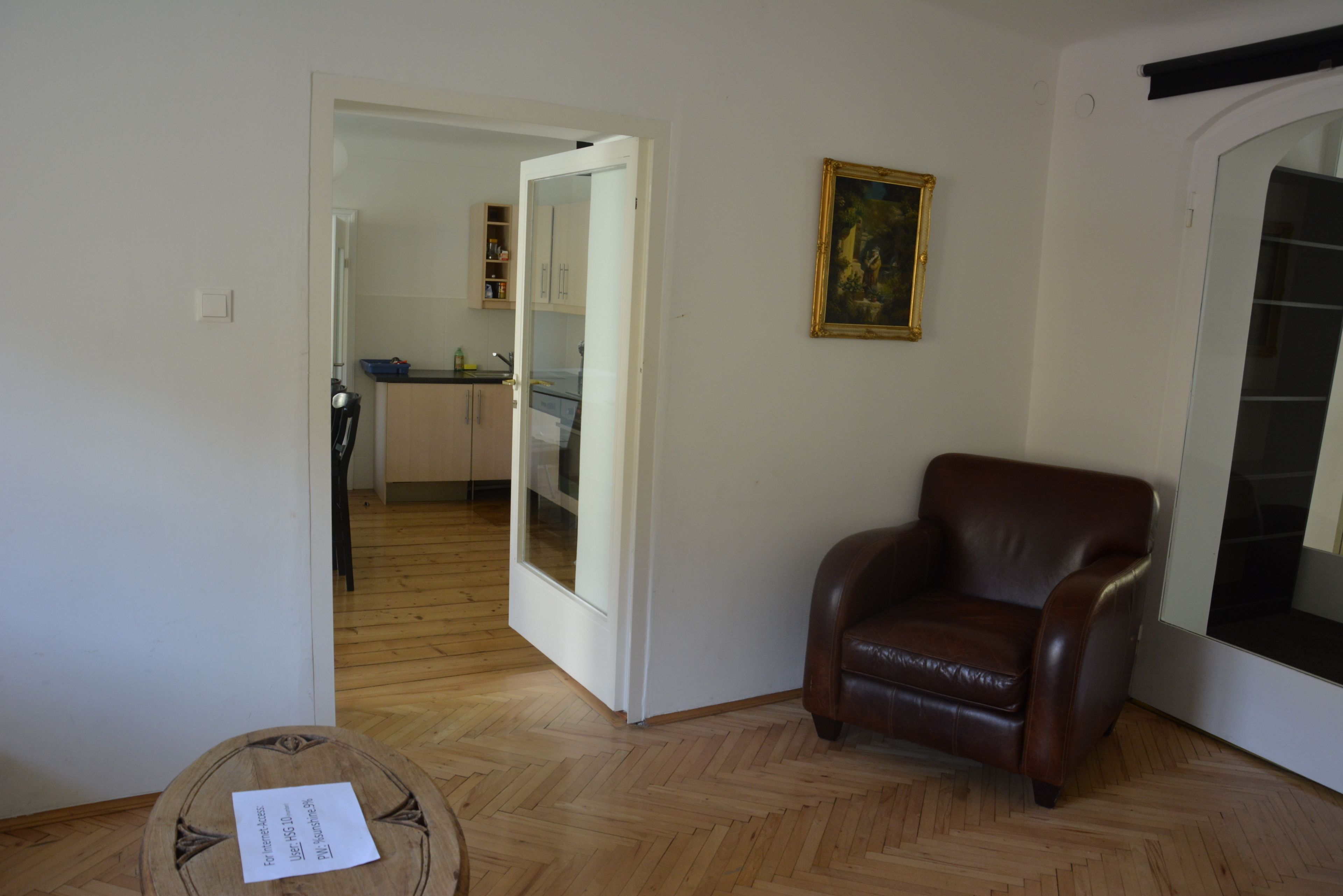 A room with a wooden herringbone floor, a brown leather armchair, a painting on the wall, and a kitchen visible through an open door.