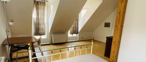 A bedroom with a wooden floor, a bed, a desk with a chair, a lamp, and a window with curtains.