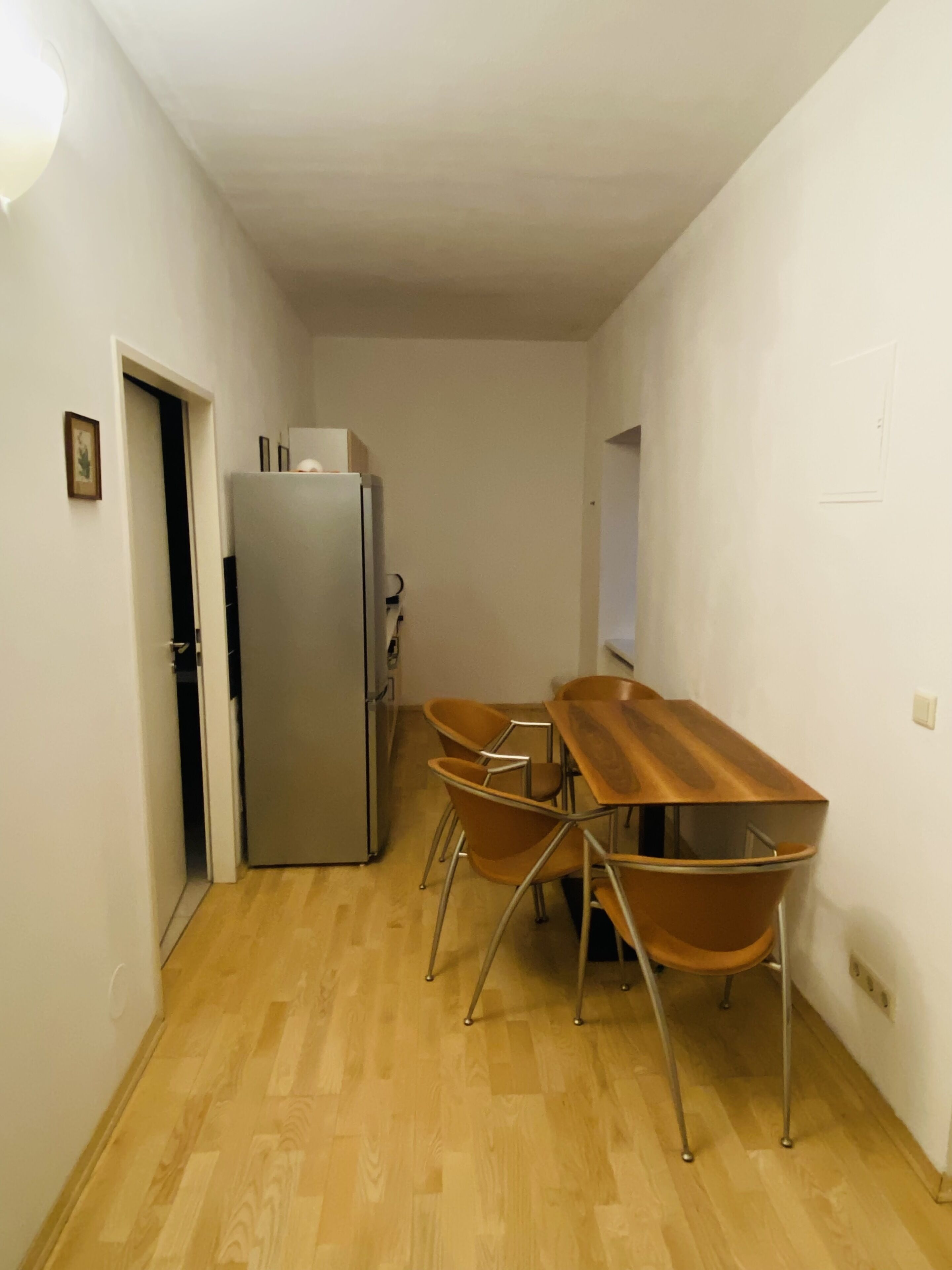Eine kleine Küche mit Holztisch und -stühlen, einem Kühlschrank und einer Tür zu einem weiteren Raum.