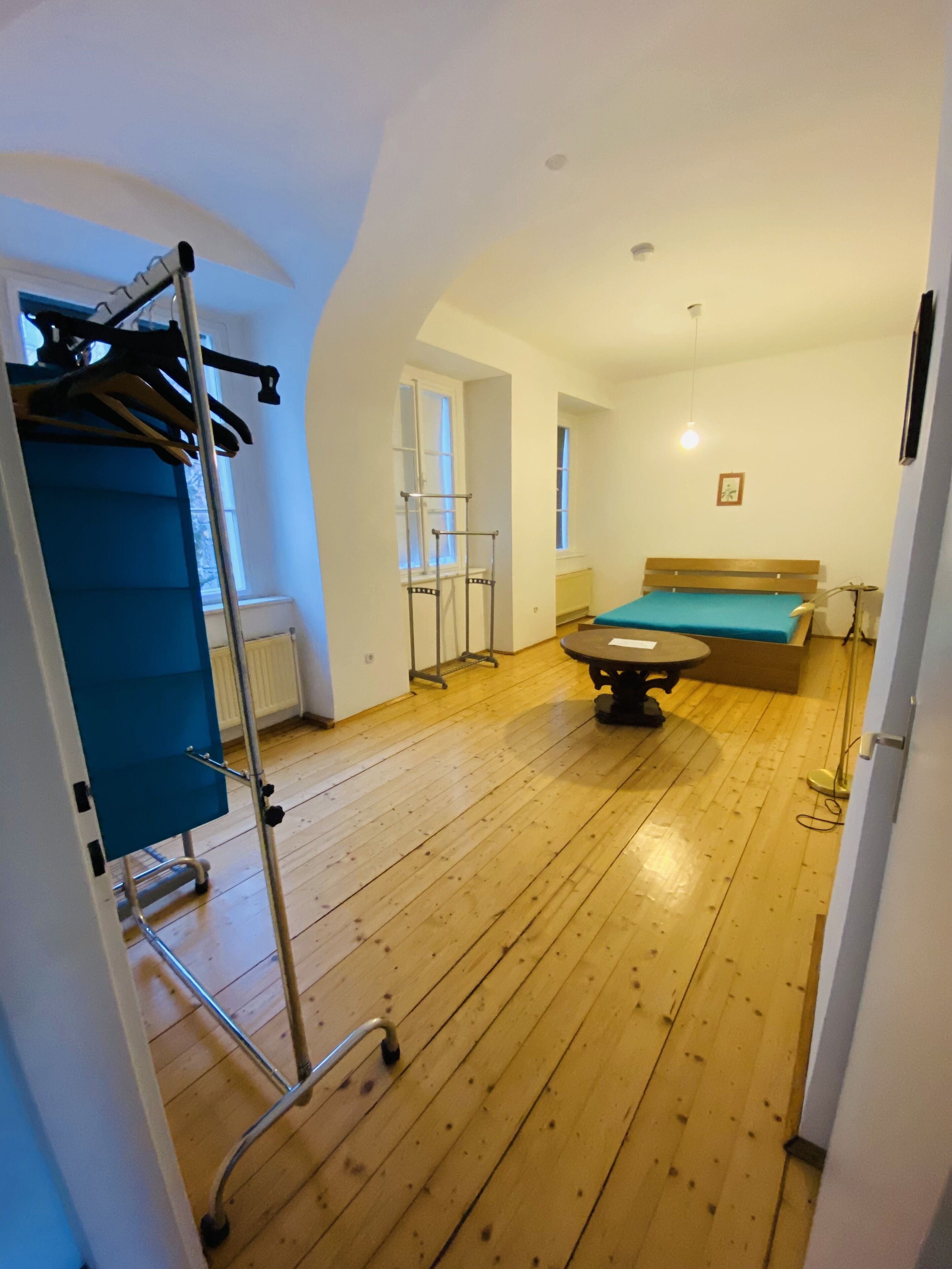 Una habitación con suelo de madera, una cama con colchón verde, una mesa, un perchero y un radiador.