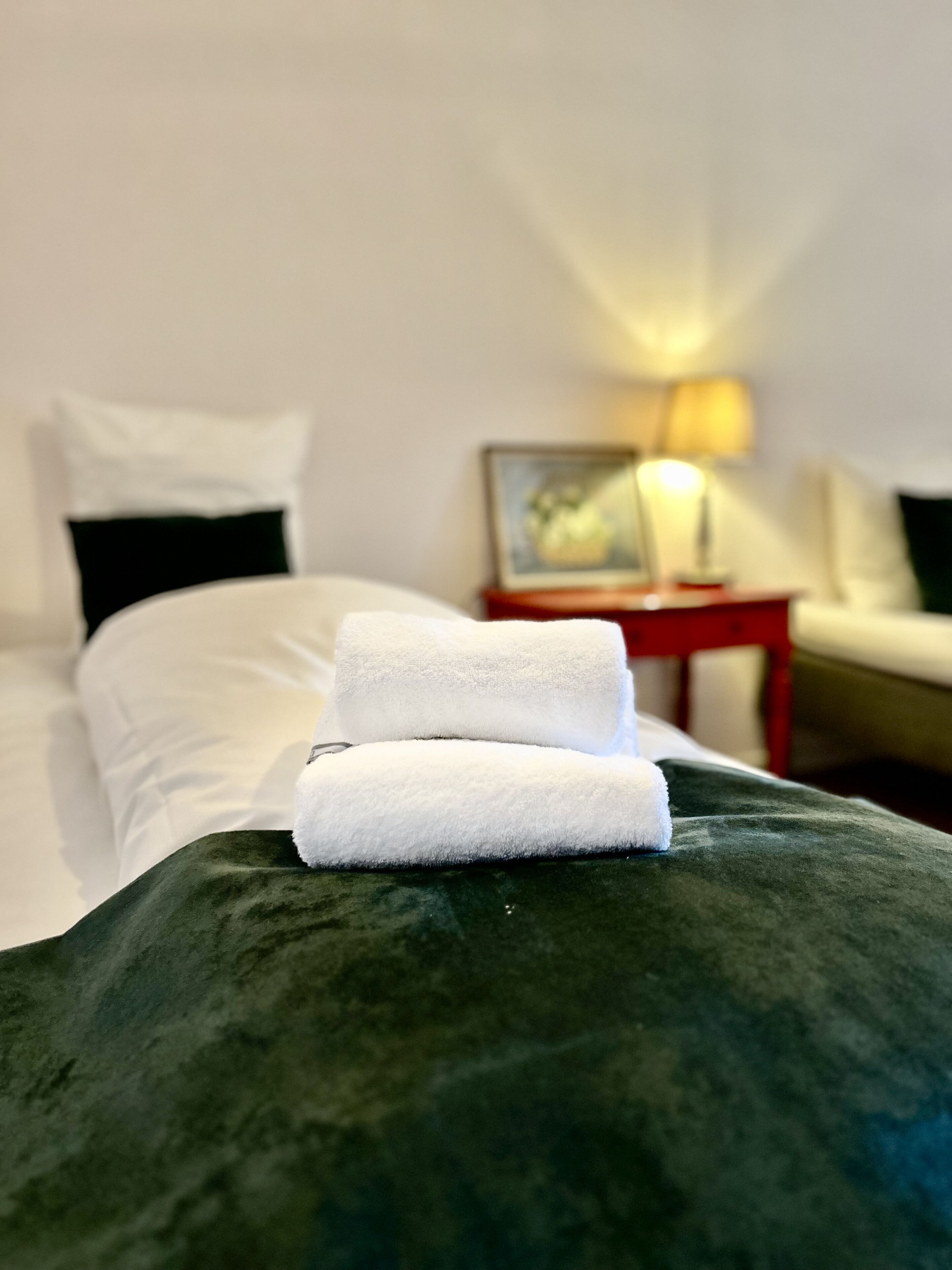 Ein ordentlich bezogenes Bett mit weißen Leinen und einem gefalteten Handtuch. Am Bett befindet sich ein roter Nachttisch mit einer Lampe und einem Bilderrahmen.