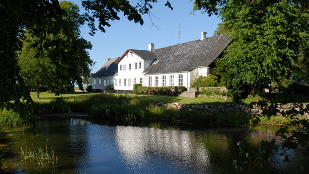 A large white building with a sloped roof, surrounded by lush greenery and a pond.