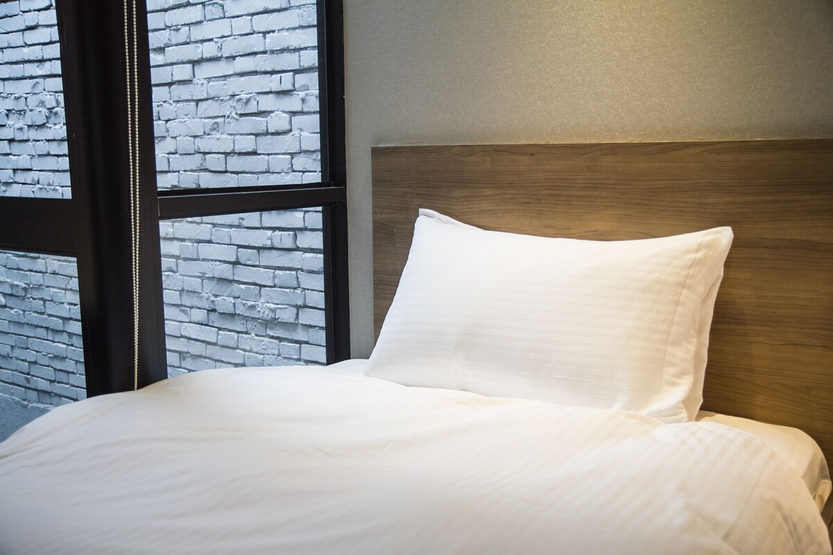 A bed with white bedding, a wooden headboard, and a brick wall visible through a window.