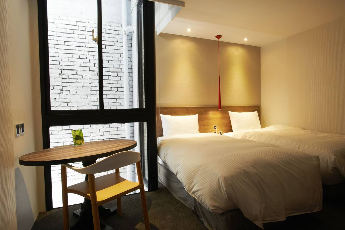 Une chambre d’hôtel avec un lit, une table et une chaise. Il y a une fenêtre donnant sur un mur en briques.