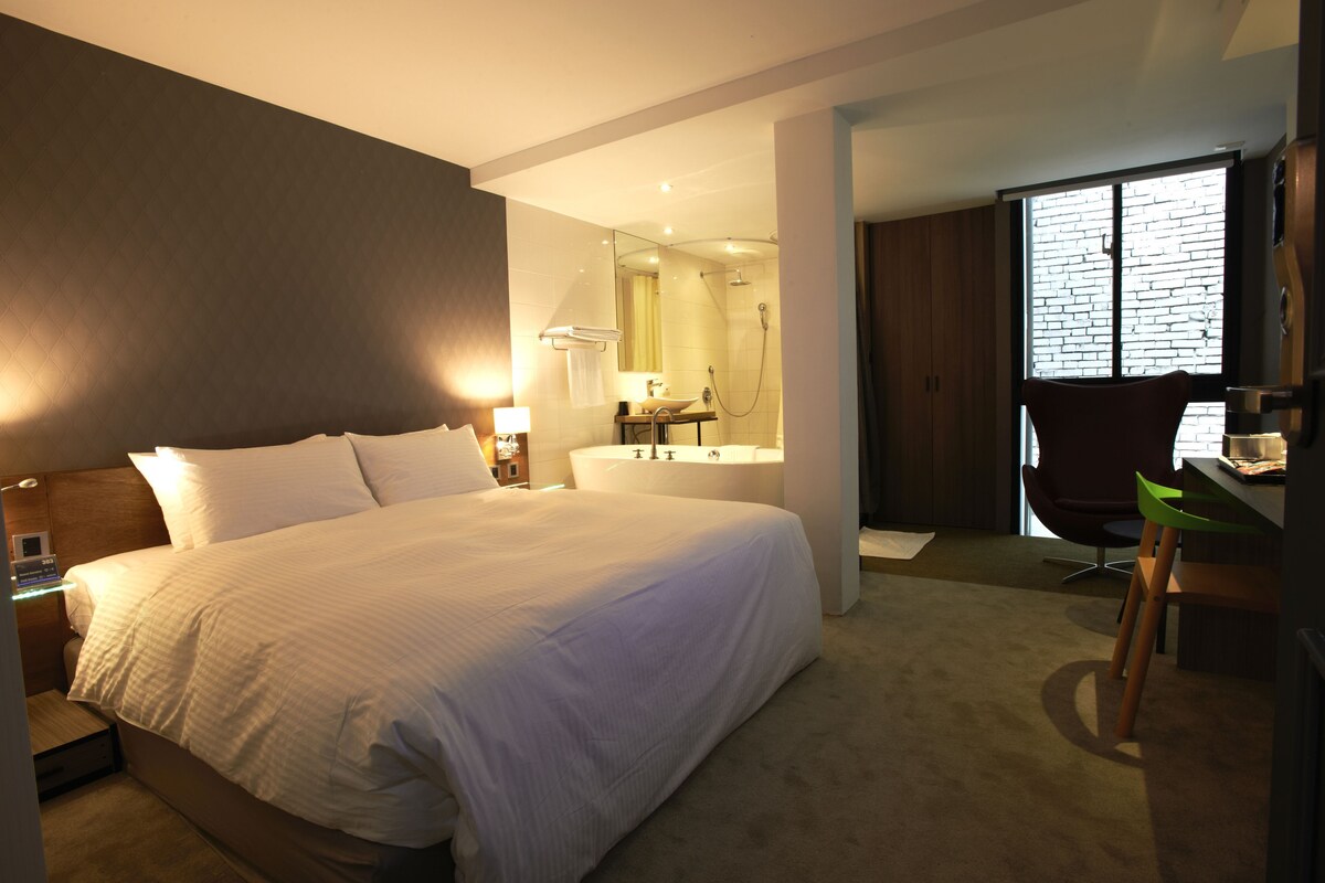 A hotel room with a large bed, a bathroom with a bathtub, a desk with a chair, and a large window.