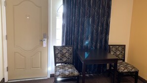 Habitación de hotel con puerta, ventana con cortinas, mesa de comedor con sillas y alfombra estampada.