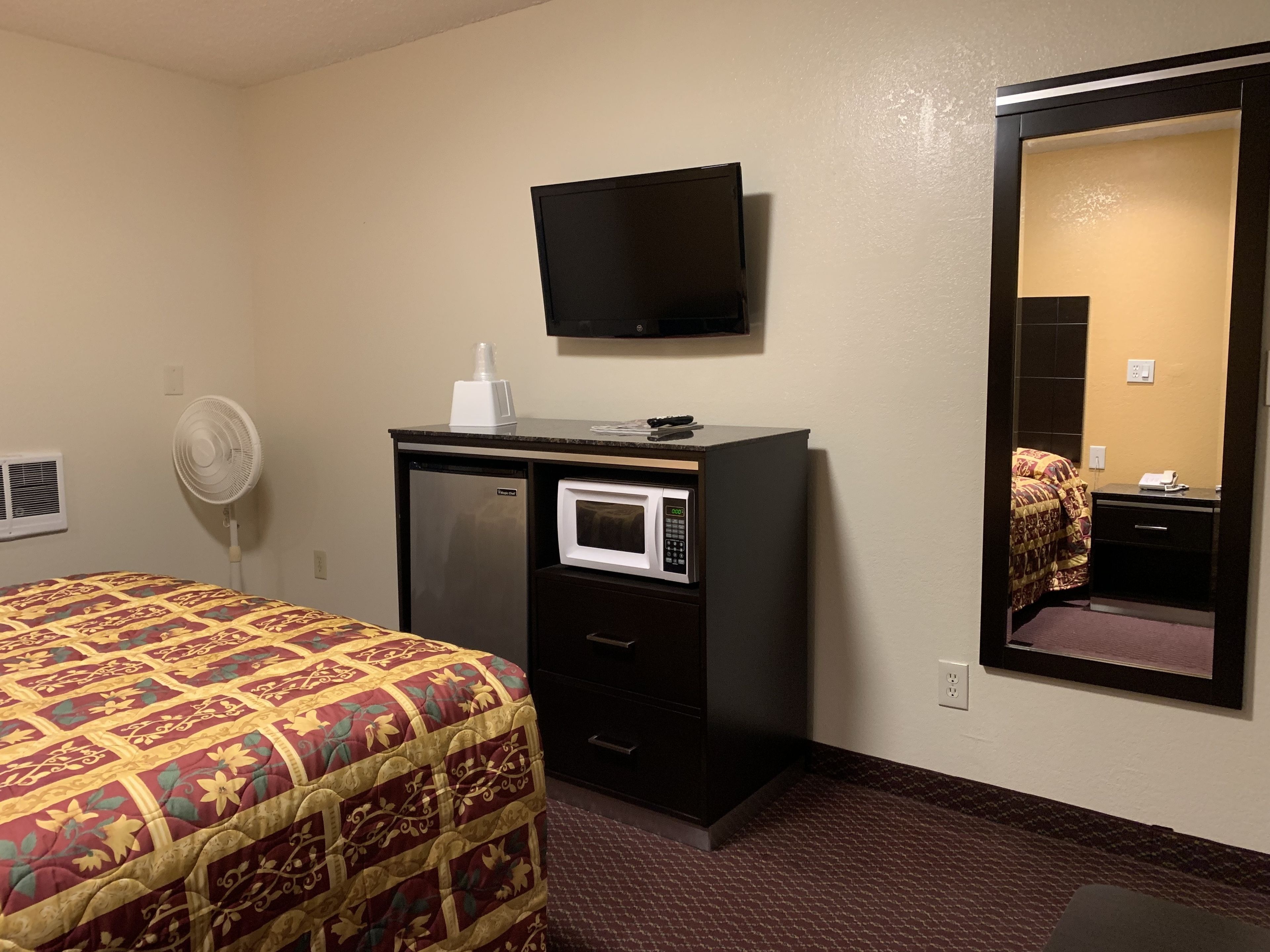 A hotel room with a bed, a desk with a microwave, a TV, and a large mirror.