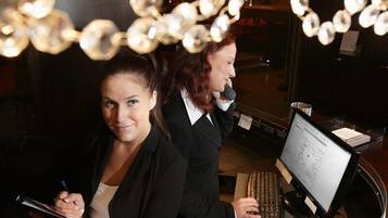 Two individuals at a desk with a computer and a notepad, in a room with a visible sign and ambient lighting.