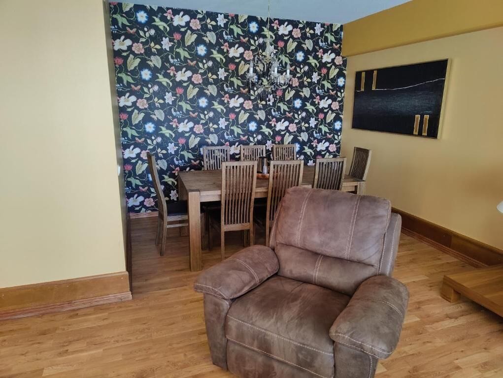A living room with a brown leather sofa, wooden dining table, and chairs. A floral wallpaper is on the wall behind the dining area.