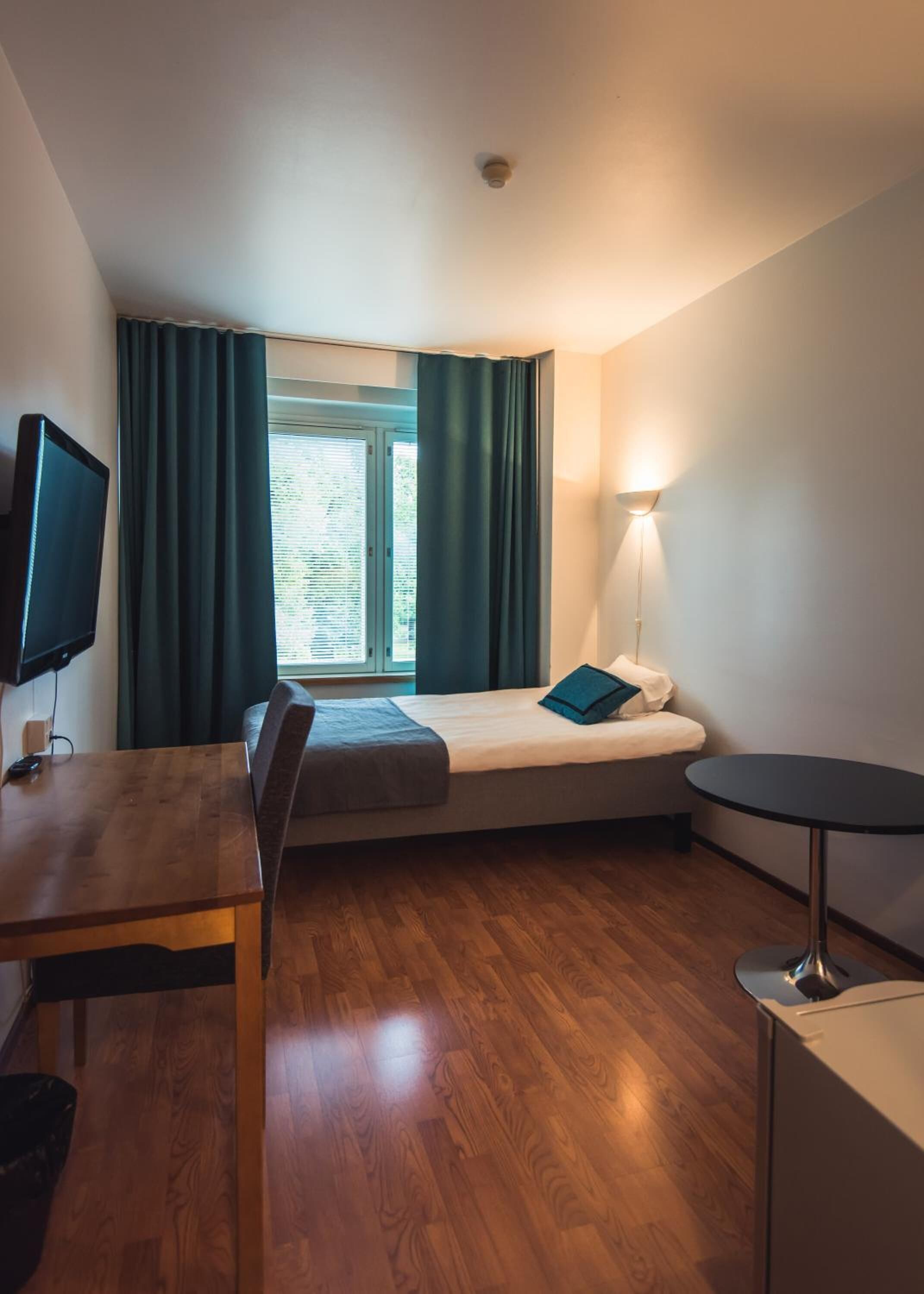 A compact hotel room with a bed, a desk, a chair, a small table, a TV, and a window with curtains.