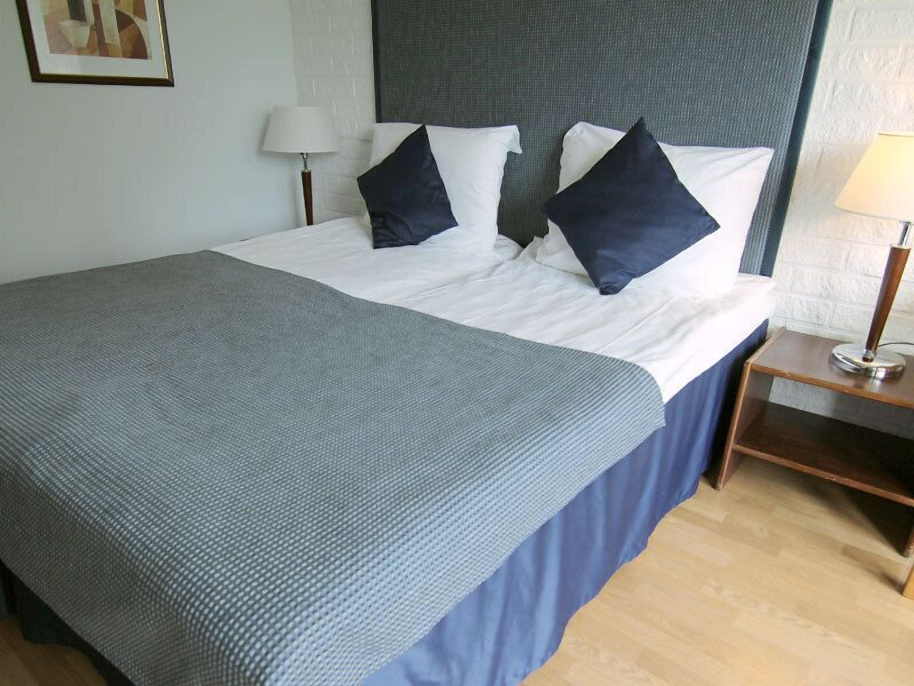 Una cama bien hecha con colcha gris y cubrecama azul, una mesita de noche con lámpara y un cuadro en la pared.