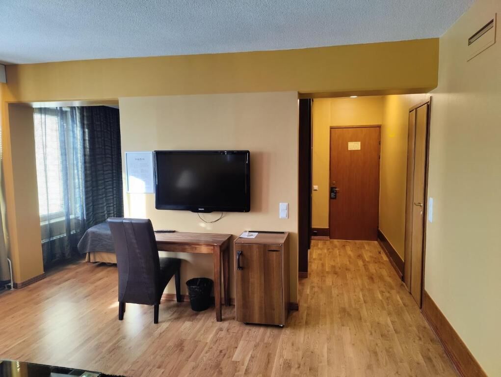 A hotel room with a wooden desk, a chair, a television mounted on the wall, and a door leading to another room.