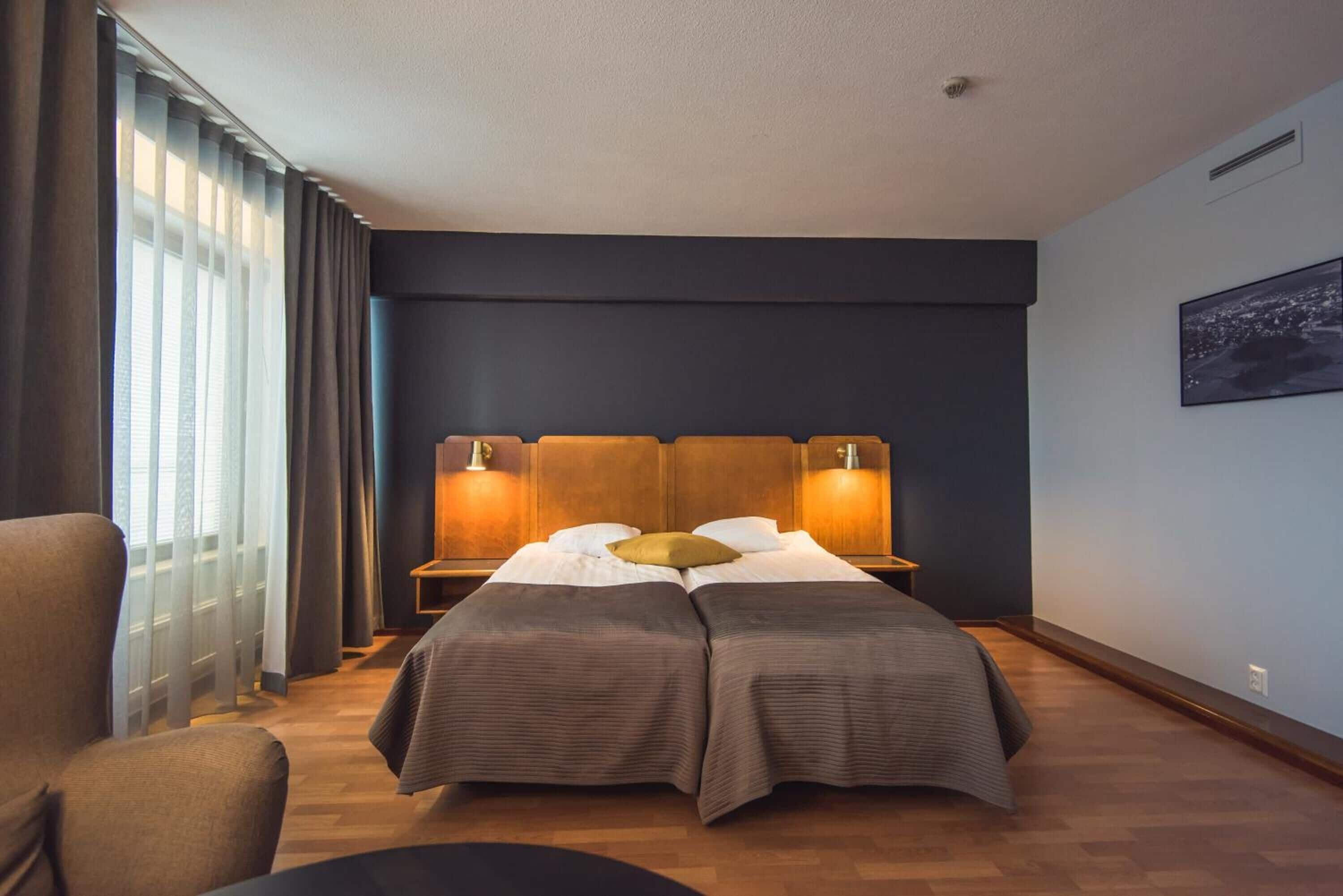Habitación de hotel con cama doble, dos mesitas de noche, una silla y un ventanal con cortinas.