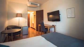 A hotel room with a bed, a desk, a chair, a small fridge, a lamp, a TV, and an air conditioner.