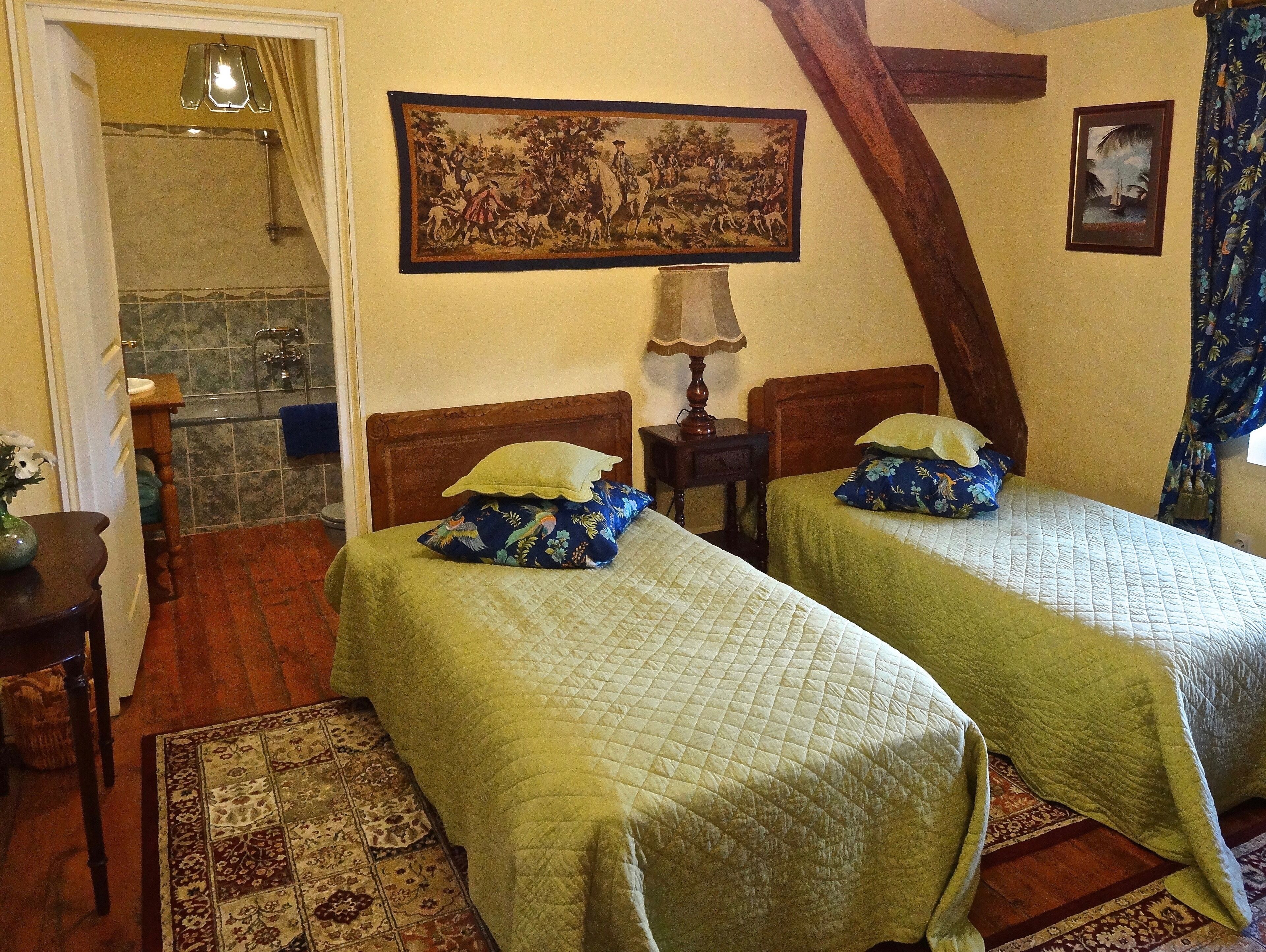 Una habitación con dos camas, cabecera de madera, un cuadro en la pared, una mesita con una lámpara y un baño visible a través de una puerta abierta.