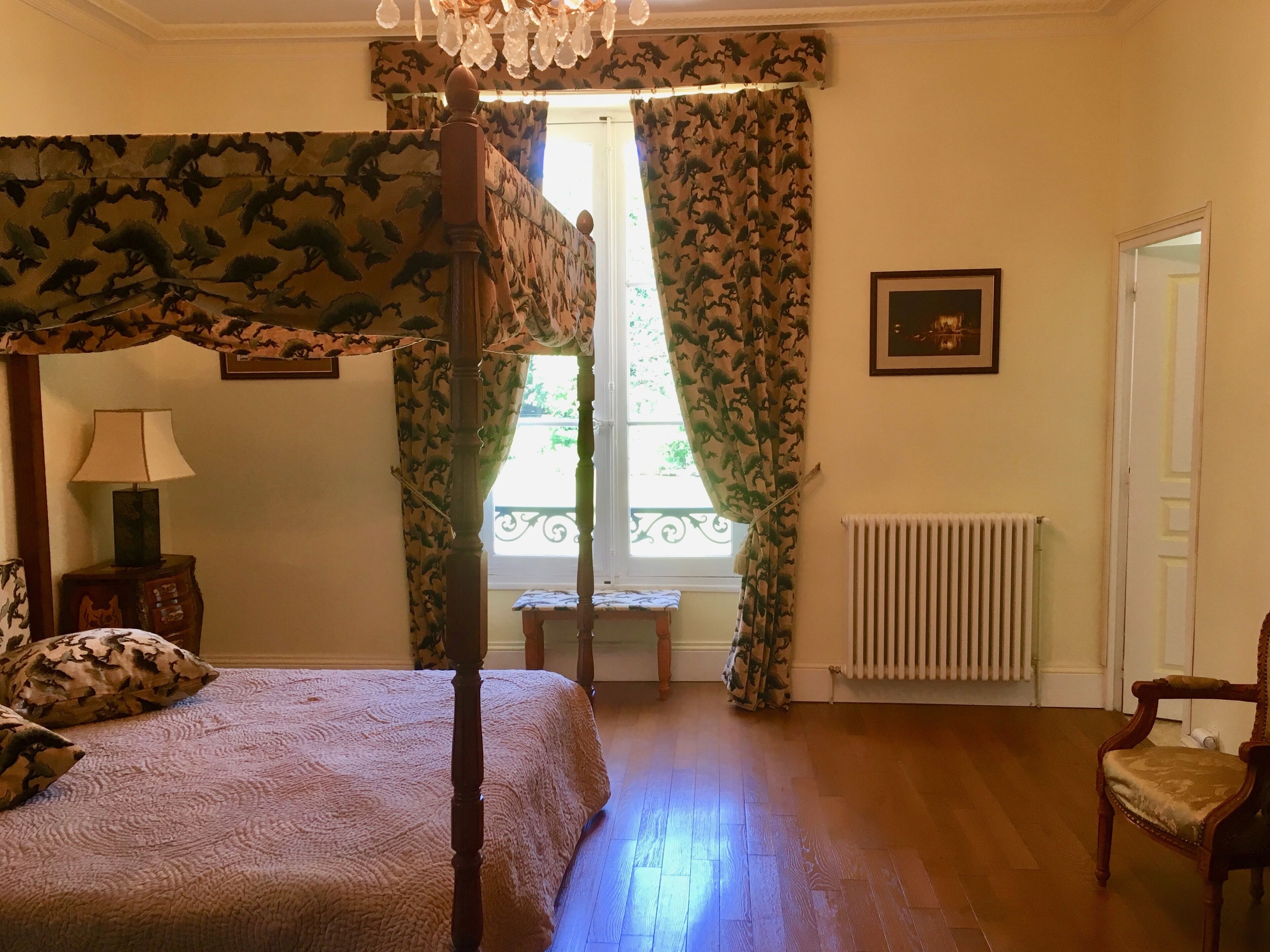 A bedroom with a four-poster bed, a chair, a radiator, and a window with curtains.